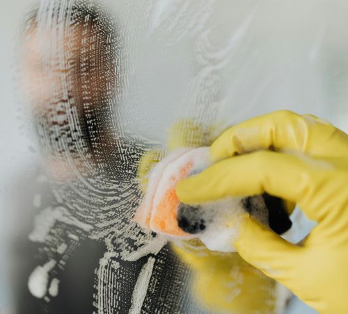 A man wearing yellow gloves cleans a bathroom mirror with a soapy sponge, ensuring hygiene and cleanliness.